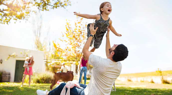 un homme assis dans l'herbe lance en l'air sa fille qui mime un oiseau